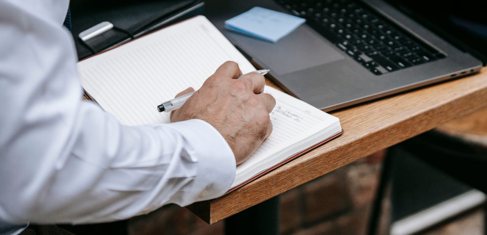 Hand writing a checklist in a notebook beside an open laptop on a desk